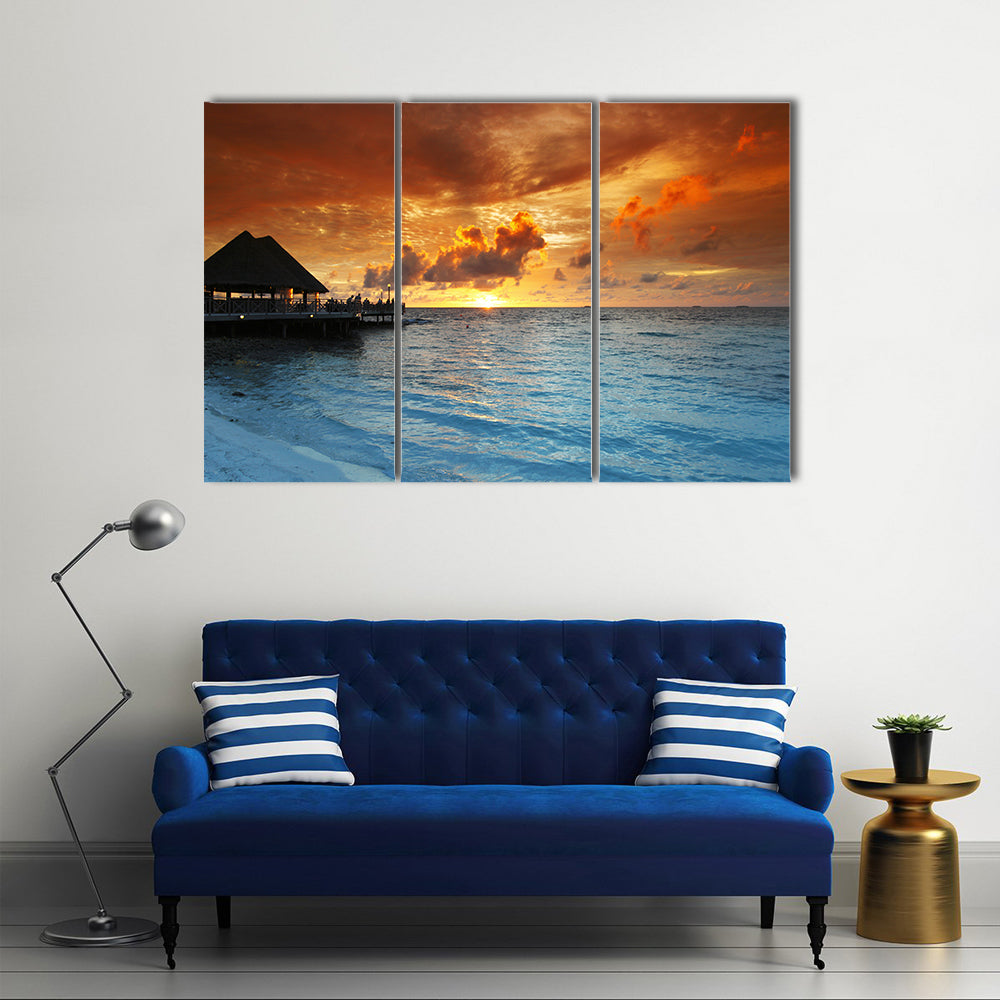 Maldives Thatched Hut on Pier at Sunset Canvas Wall Art in orange, yellow and blue, 3 panel horizontal set hung on a wall
