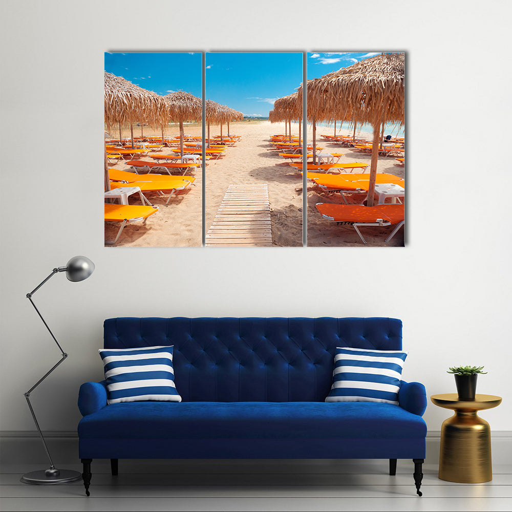 Greek Beach with Wooden Walkway and Orange Loungers Canvas Wall Art in beige, orange and blue, 3 panel horizontal set hung on a wall
