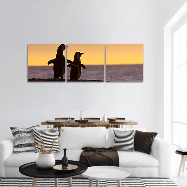 Two Penguin Silhouettes at Golden Sunset Panoramic Canvas Wall Art in black, orange and yellow, hung on a wall