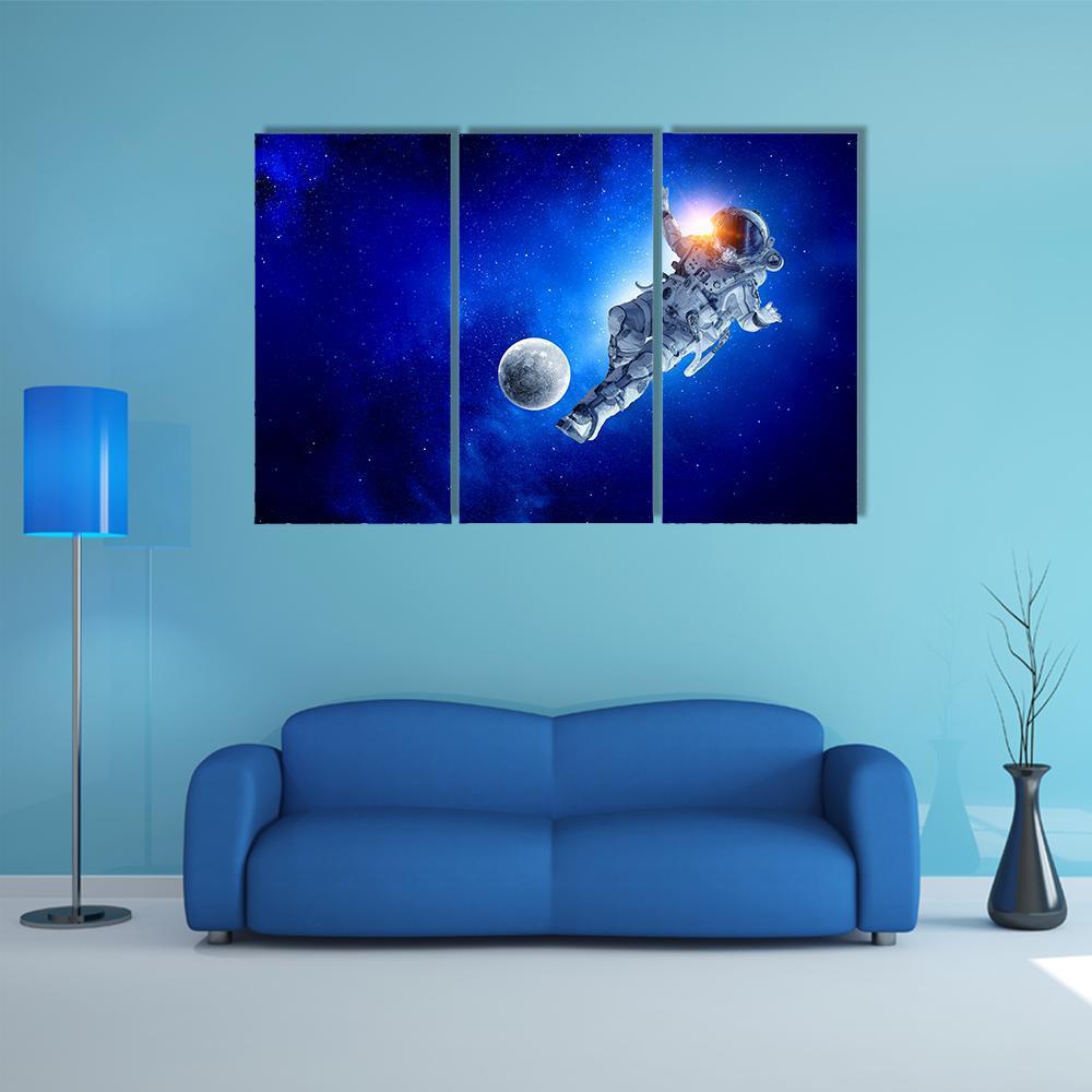 Astronaut Kicking Glowing Ball Near Moon Canvas Wall Art in black, white and blue, 3 panel horizontal set hung on a wall