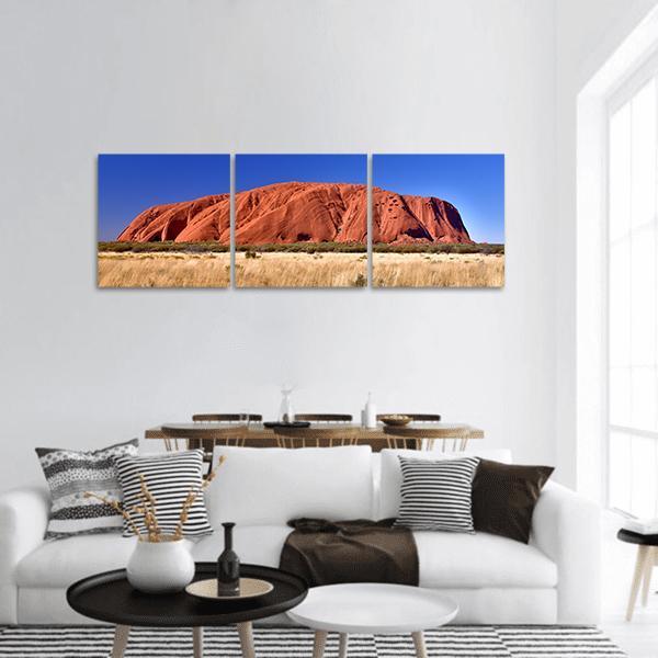 Ayers Rock in Sunlit Outback Panoramic Canvas Wall Art in red, orange and blue, hung on a wall