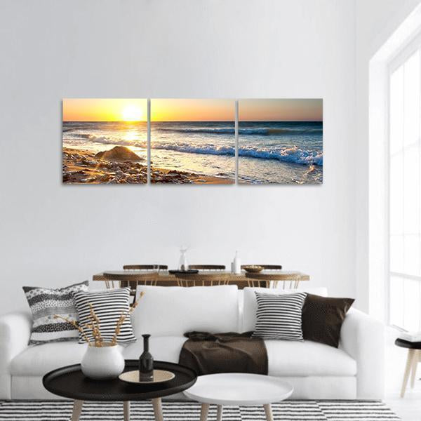 Sundown Seascape Over Rocky Shore Panoramic Canvas Wall Art in orange, yellow and blue, hung on a wall