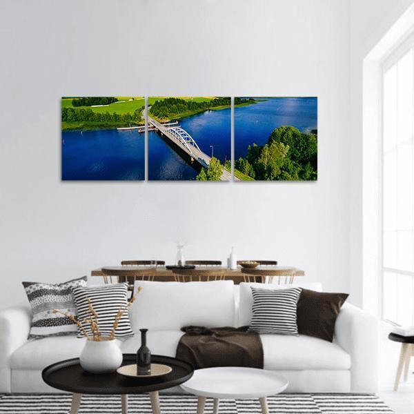 White Arched Bridge Over Blue Lake Panoramic Canvas Wall Art in green and blue, hung on a wall