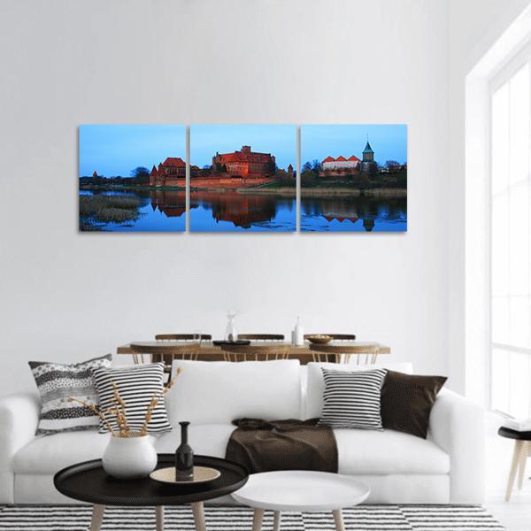 Castle of the Teutonic Knights at Lake Dusia Panoramic Canvas Wall Art in brown, orange and blue, hung on a wall