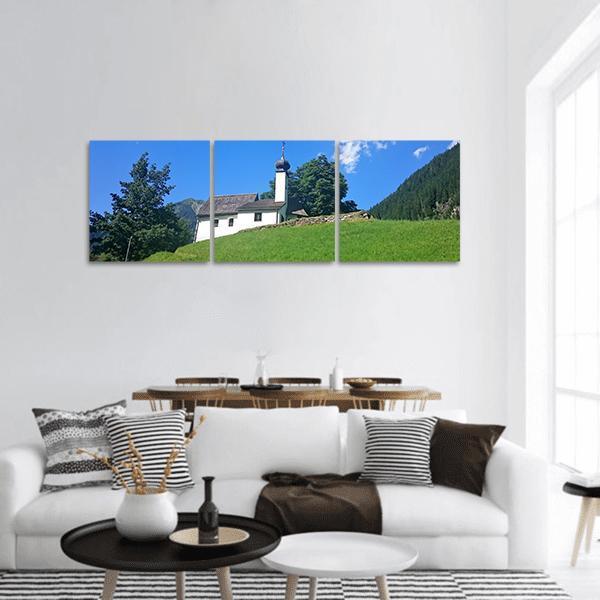 White Alpine Church on Hilltop Panoramic Canvas Wall Art in white, green and blue, hung on a wall