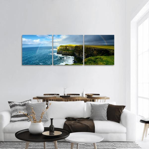 Rainbow Over Coastal Cliffs Panoramic Canvas Wall Art in green and blue, hung on a wall