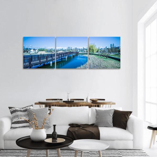 Wooden Bridge Over Willow-lined Mountain Lake Panoramic Canvas Wall Art in brown, green and blue, hung on a wall