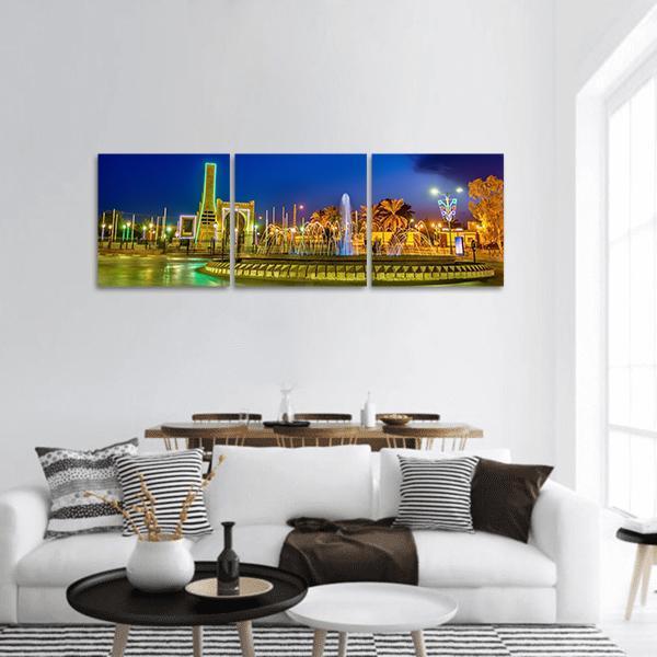 Illuminated Fountain and Tower at Night Panoramic Canvas Wall Art in yellow, green and blue, hung on a wall