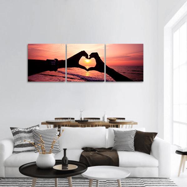 Hands Forming Heart at Sunset Panoramic Canvas Wall Art in black, orange and pink, hung on a wall