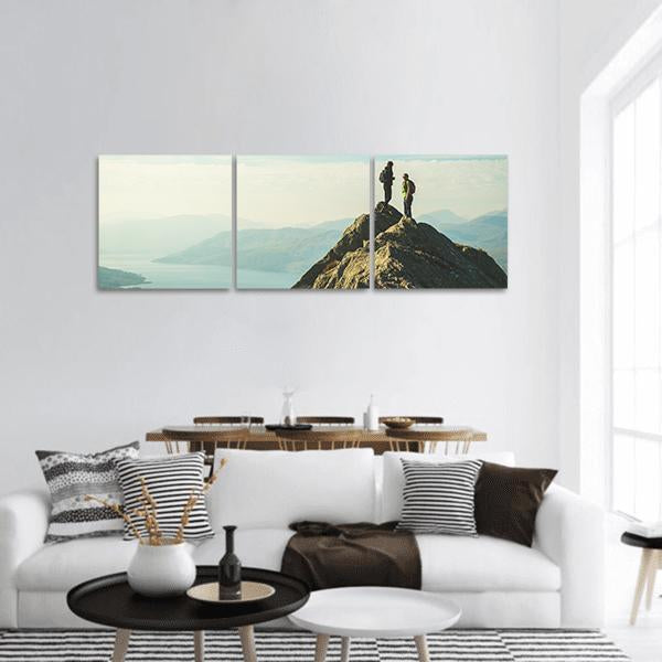 Hikers on Mountain Summit Over Lake Panoramic Canvas Wall Art in white, gray and blue, hung on a wall