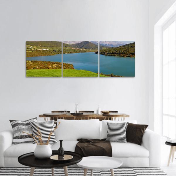 Calm Mountain Lake at Distant Peaks Panoramic Canvas Wall Art in brown, green and blue, hung on a wall