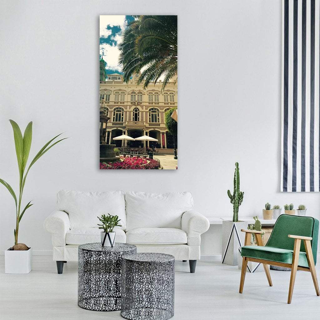 Spanish Plaza Fountain with Pink Flowers Vertical Canvas Wall Art in beige, green and blue, single vertical panel hung on a wall