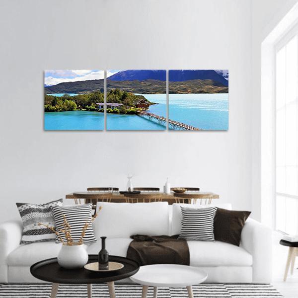 Island with Wooden Footbridge Over Turquoise Lake Panoramic Canvas Wall Art in brown, green and blue, hung on a wall