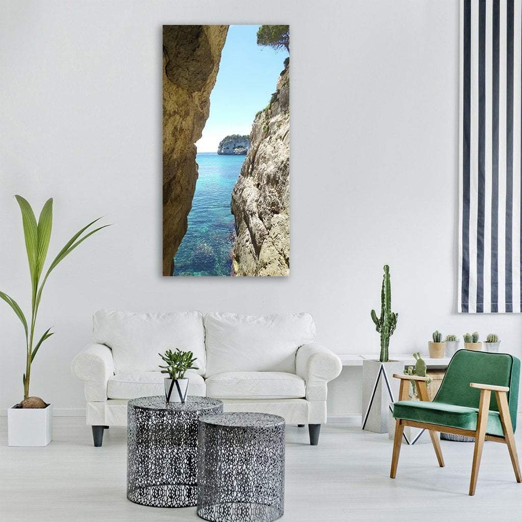 Menorca Sea Passage Between Limestone Cliffs Vertical Canvas Wall Art in beige, brown and blue, single vertical panel hung on a wall