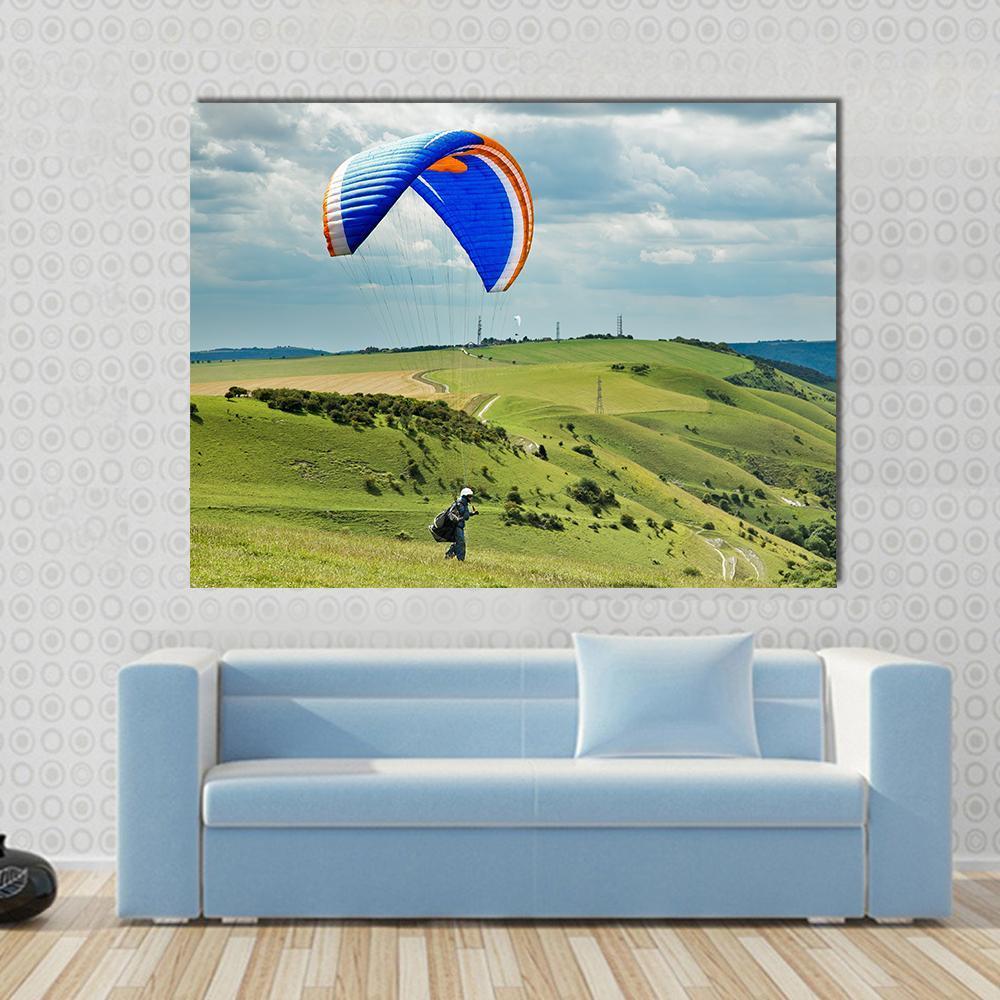 Blue and Orange Paraglider Over Rolling Green Hills Canvas Wall Art in white, green and blue, single panel hung on a wall