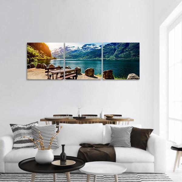 Bench by Lake with Snow-capped Mountains Panoramic Canvas Wall Art in white, green and blue, hung on a wall