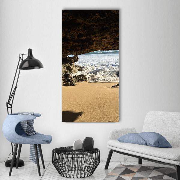 Beach Through Rock Archway Vertical Canvas Wall Art in beige, brown and blue, single vertical panel hung on a wall