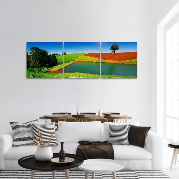 Red-earth Hill with Lone Tree and Lake Panoramic Canvas Wall Art in red, green and blue, hung on a wall