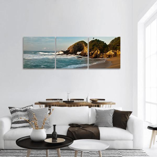 Sandy Beach with Rocks at Sunrise Panoramic Canvas Wall Art in brown, gold and blue, hung on a wall