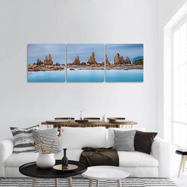 Jagged Sea Stacks on Calm Blue Coast Panoramic Canvas Wall Art in gray, brown and blue, hung on a wall