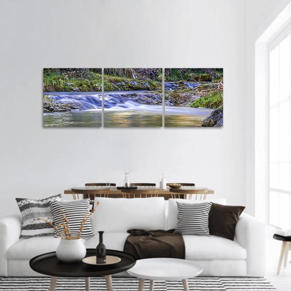 Silky Forest Stream with Small Waterfall Panoramic Canvas Wall Art in brown, green and blue, hung on a wall