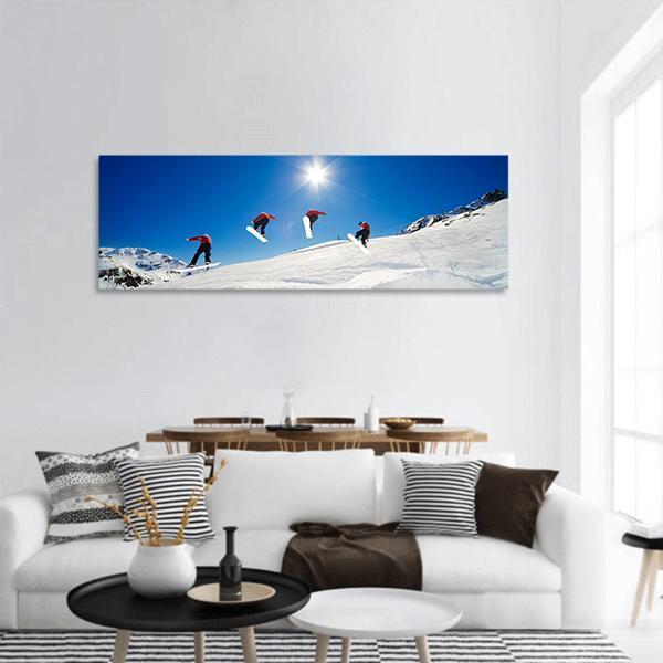 Four Snowboarders Mid-air Over Snow Slopes Panoramic Canvas Wall Art in white and blue, single panel hung on a wall