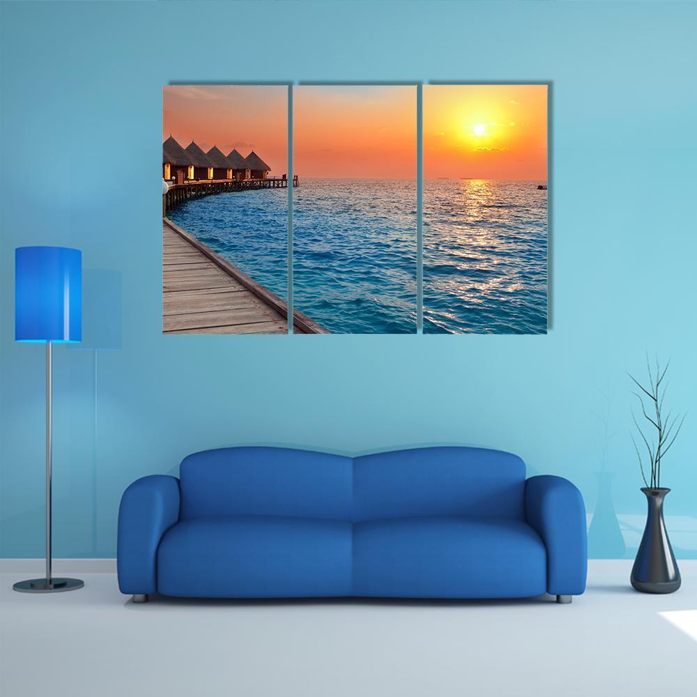 Sunset Over Overwater Bungalows and Pier Canvas Wall Art in orange, yellow and blue, 3 panel horizontal set hung on a wall