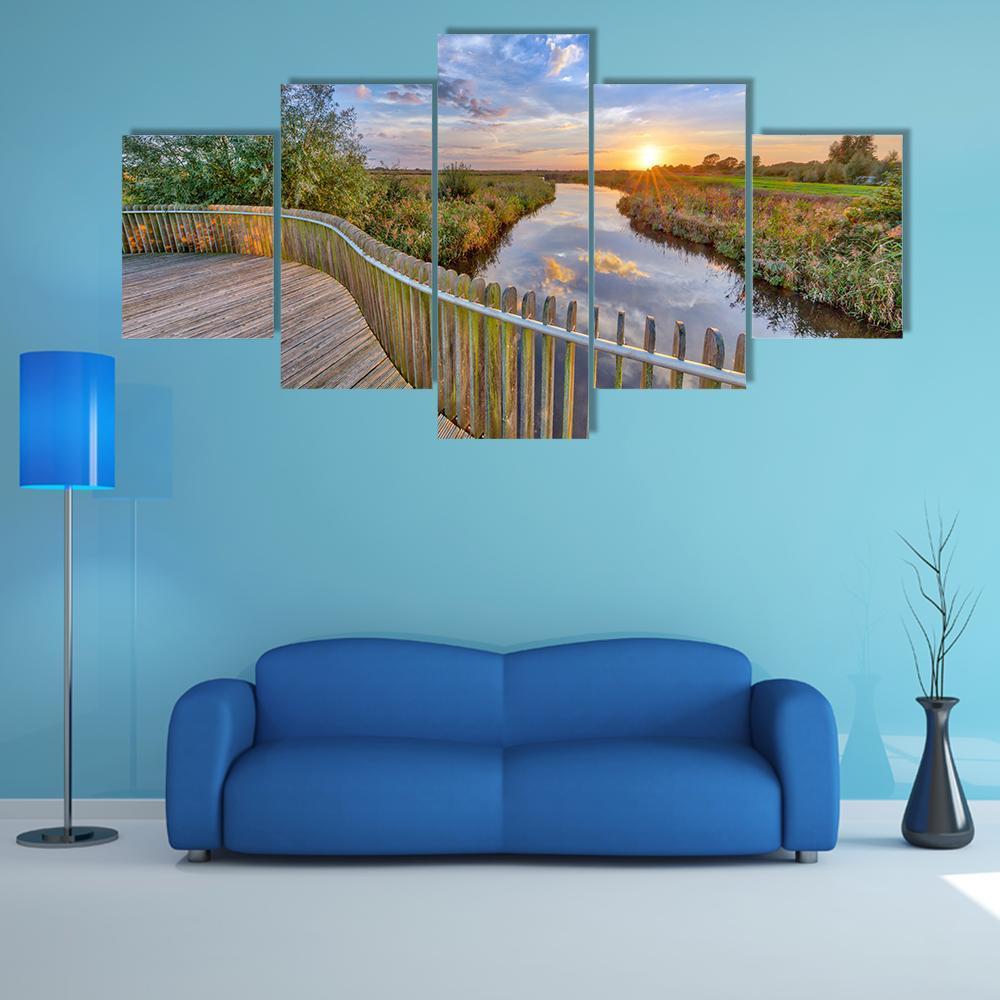 Sunset Over Wooden Boardwalk and River Canvas Wall Art in brown, orange and blue, 5 panel Star layout hung on a wall