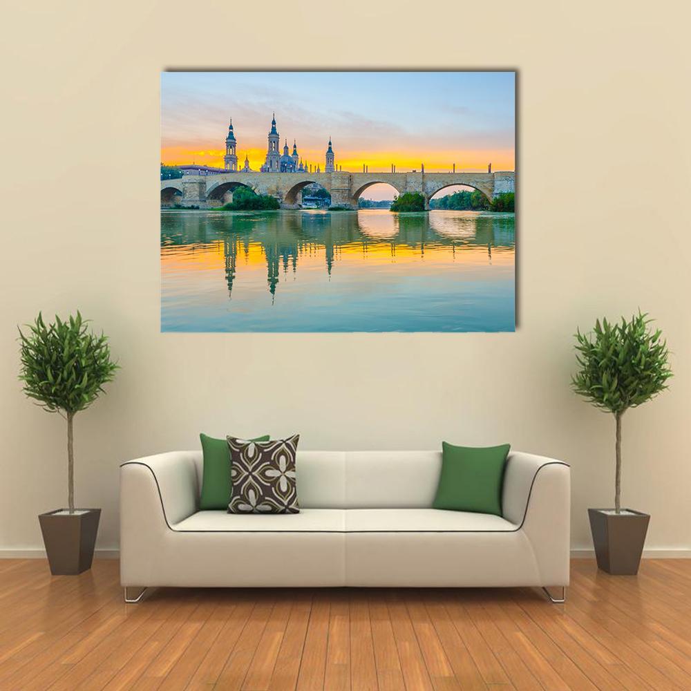 Sunset Basilica Over River and Stone Arch Bridge Canvas Wall Art in orange, yellow and blue, single panel hung on a wall