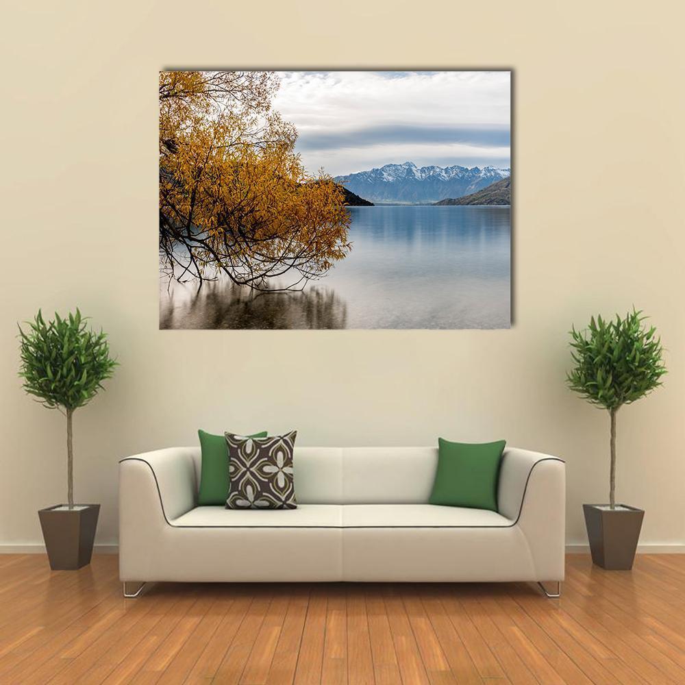 Tree-framed Lake with Snow-capped Mountains Canvas Wall Art in gray, yellow and blue, single panel hung on a wall