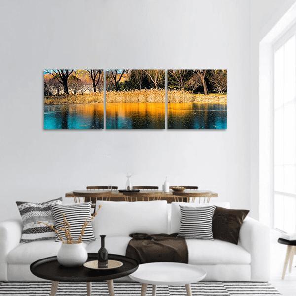 Autumn Trees by the Lake Panoramic Canvas Wall Art in orange, yellow and blue, hung on a wall