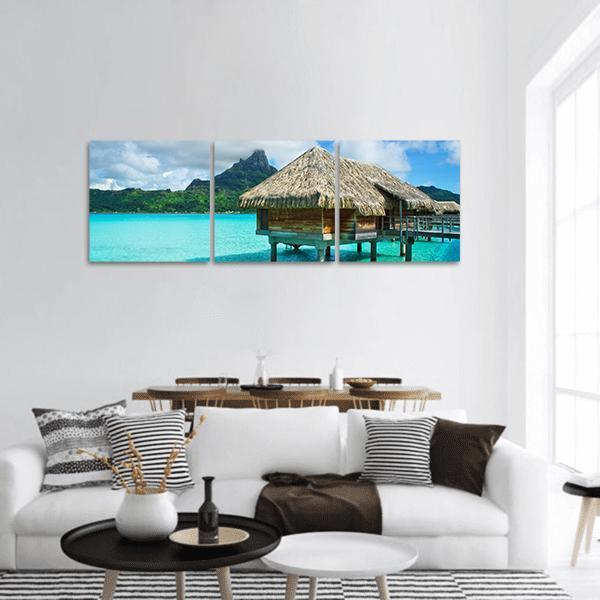 Thatched Overwater Bungalow in Turquoise Lagoon Panoramic Canvas Wall Art in brown, green and blue, hung on a wall