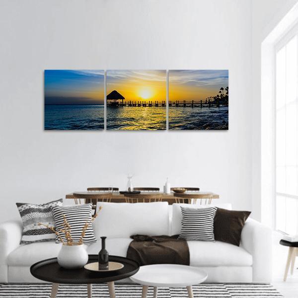 Tropical Sunset Over Thatched Pier Panoramic Canvas Wall Art in orange, yellow and blue, hung on a wall