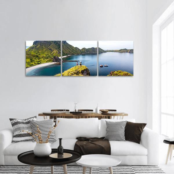 Hikers on Grassy Cliff Overlooking Coastal Bay Panoramic Canvas Wall Art in green and blue, hung on a wall