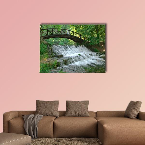 Wooden Arched Bridge Over Tiered Waterfall Canvas Wall Art in white, brown and green, single panel hung on a wall